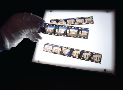 Gloved hand holding strip of negatives over lightbox. Two strips of negatives rest on lightbox.